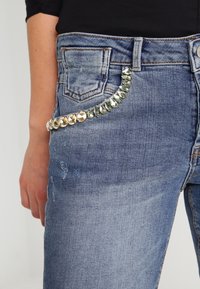 Primer plano de unos vaqueros de denim azul con detalles desgastados y una cadena de piedras brillantes acoplada al bolsillo delantero, usados por una persona con una camiseta negra.