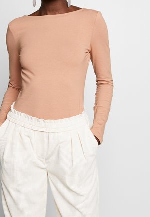 Personne portant un haut beige à manches longues avec un pantalon en velours côtelé blanc, taille haute et plissé, et des boucles d'oreilles blanches pendantes, les mains dans les poches.