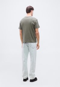 Άνδρας που στέκεται με την πλάτη γυρισμένη, φοράει λαδί κοντομάνικο πουκάμισο, ανοιχτόχρωμα τζιν και μαύρα παπούτσια σε λευκό απλό φόντο.