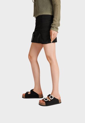 Donna che indossa una minigonna nera arricciata, un maglione di lana verde oliva e sandali neri aperti con grandi fibbie quadrate d'argento.