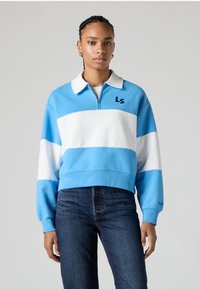 Sudadera corta azul claro con rayas horizontales blancas, cuello con cremallera y logo bordado "LS", combinada con vaqueros oscuros.