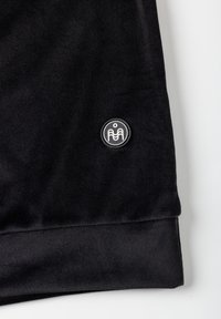 Musta corduroy-collegepaita, jossa pyöreä metallinen logopatch vasemmassa hihassa ja teksturoidut ribbiresorit.