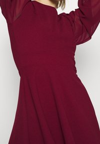 Kvinna i en ärmlös bordeauxfärgad klänning med genomskinliga långa ärmar, upplyfta armar och en figursydd midja mot en enfärgad bakgrund.