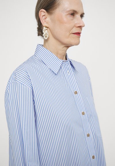 Blue and white striped button-up shirt with a classic collar and wooden buttons. Model wearing decorative pearl hoop earrings.