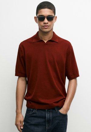 Jonge man met zwarte zonnebril, een donkerrode polo met korte mouwen en donkere spijkerbroek met één hand in de zak tegen een witte achtergrond.