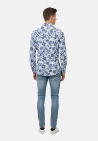 Hombre de espaldas que lleva una camisa blanca de manga larga con estampado floral azul, jeans azul claro y zapatillas blancas con detalles negros en el talón.