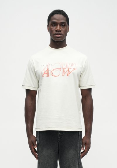 Camiseta de color beige claro con texto gráfico rojo en negrita en la parte frontal, mangas cortas y un ajuste holgado. Material de algodón con costuras visibles.