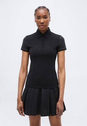 Γυναίκα που φοράει εφαρμοστή μαύρη πόλο μπλούζα με κοντά μανίκια και κουμπιά, καθώς και μαύρη πλισέ φούστα, στέκεται μπροστά σε ένα απλό ανοιχτό φόντο.