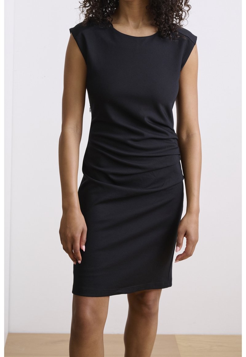 Femme portant une robe noire moulante sans manches, longueur genou, debout contre un mur blanc uni et un sol en bois.