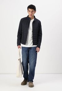 Giacca nera trapuntata con colletto alto, indossata sopra una camicia bianca, abbinata a jeans blu e scarpe beige. Tiene in mano una borsa a tracolla beige.