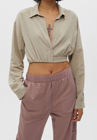 Chemise beige raccourcie avec un col en V profond et des manches longues, associée à un pantalon rose pâle à taille élastique avec poches latérales.
