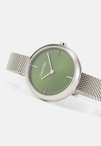 Reloj con correa de malla plateada, esfera redonda verde, marcadores de hora plateados minimalistas y logotipo. Presenta una caja suave y pulida.