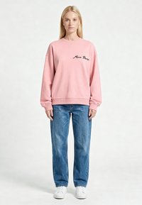 Femme debout, face à l'objectif, portant un sweat rose avec le texte « Mina Paris », un jean bleu et des baskets blanches sur un fond uni.