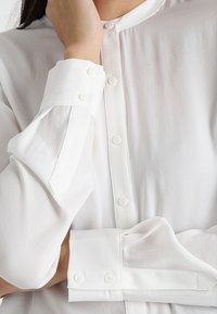 Bílá dlouhá košile s knoflíkovým zapínáním v přední části. Má vysoký límec a mírně průhlednou tkaninu. Knoflíkové detaily na manžetách rukávů.