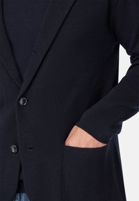 Námornícky pletený blazer s klopou, prednou vreckom, dvoma gombíkmi a pružnými manžetami, ktorý predvádza textúrovanú tkaninu.