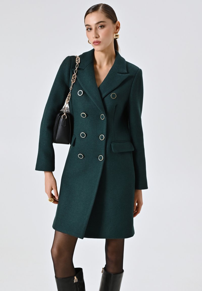 Donkergroene wollen mix jas met een dubbelzijdig ontwerp, metalen oogaccenten en gesneden revers. Gestyled met een zwarte handtas en kniehoge laarzen.