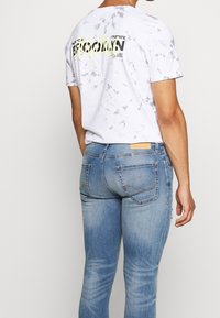 Vit tie-dye t-shirt med grafisk design där det står "Brooklyn" i gult på baksidan, tillsammans med ljusblå tajta jeans med bakfickor.