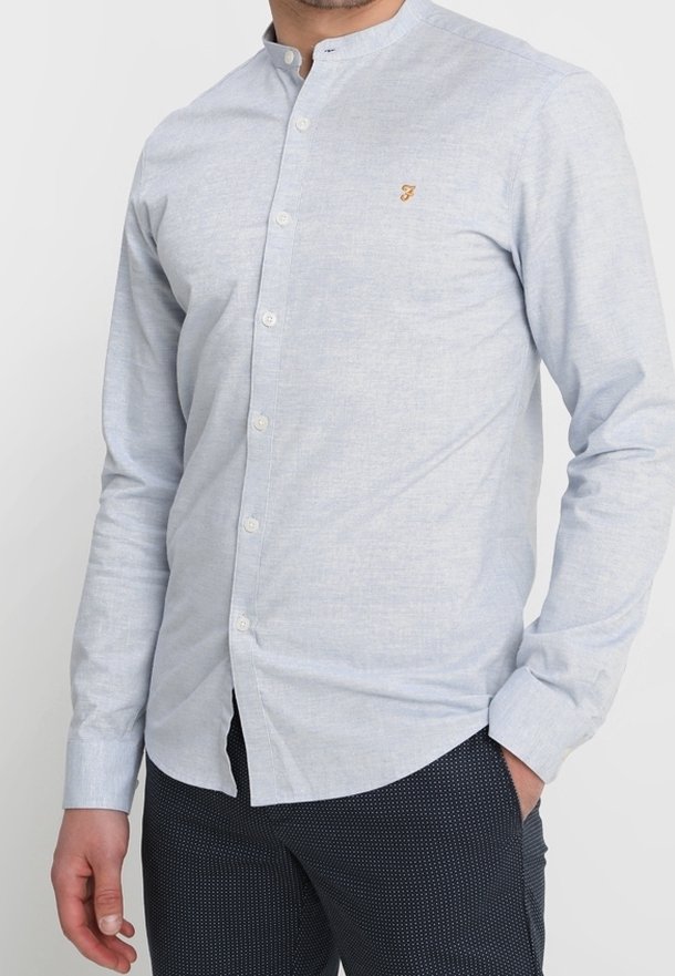 Homme portant une chemise bleu clair à boutons avec un petit logo brodé sur la poitrine et un pantalon foncé à motifs, une main dans la poche.