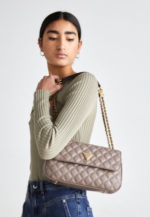 Frau trägt ein beiges, geripptes Langarmshirt und blaue Jeans, hält eine beige gesteppte Handtasche mit goldfarbenem Kettenriemen über der Schulter.