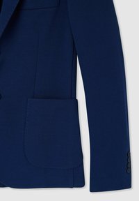 Manica di una giacca blu navy con tessuto testurizzato, una grande tasca applicata e quattro bottoni scuri al polsino, dal taglio sartoriale.