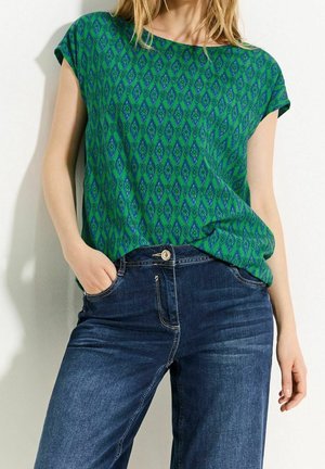 Kvinde iført grøn mønstret kortærmet top stoppet ned i mørkeblå jeans, stående med den ene hånd i lommen mod en hvid væg.