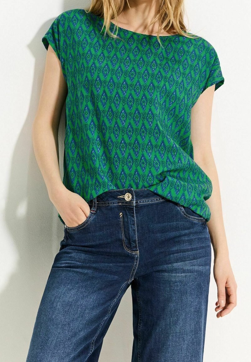 Femme portant un haut vert à manches courtes à motifs, rentré dans un jean bleu foncé, debout avec une main dans la poche contre un mur blanc.