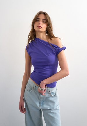 Jeune femme aux longs cheveux portant un haut violet asymétrique et un jean bleu clair, debout devant un fond blanc uni.