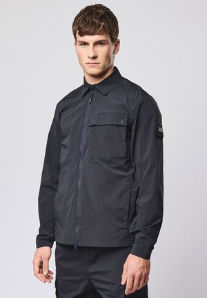 Giovane uomo che indossa una giacca nera con cerniera, una tasca con pattina sul petto e una toppa sulla manica sinistra, in piedi davanti a uno sfondo semplice.