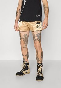 Calções de cetim dourados com logo "Venum" preto, combinados com botas de tornozelo pretas e castanhas. Tatuagens expostas apresentam desenhos intrincados nas pernas.