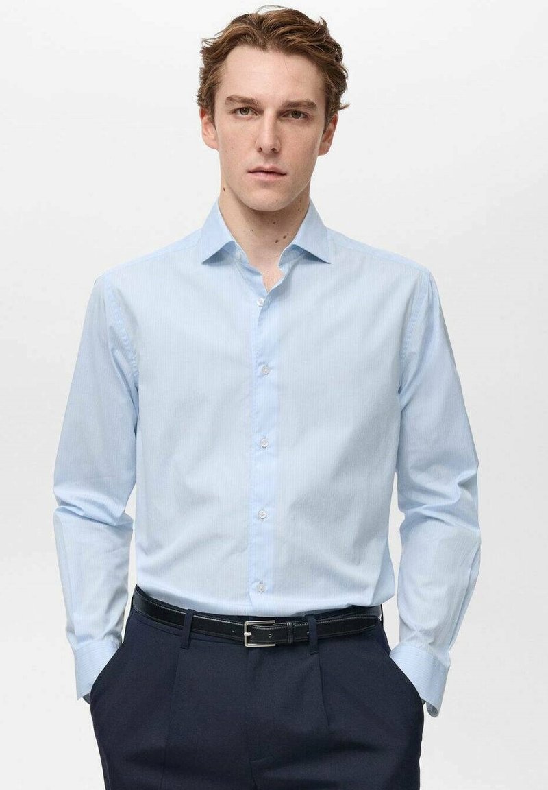 Camicia a righe blu chiaro con colletto a punta, maniche lunghe e pattina dei bottoni, abbinata a pantaloni scuri e una cintura nera.