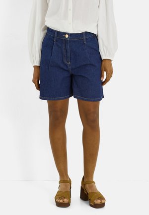 Persona que lleva puestos unos pantalones cortos vaqueros azul oscuro, una blusa blanca de manga larga y sandalias de plataforma marrones, de pie contra un fondo blanco.