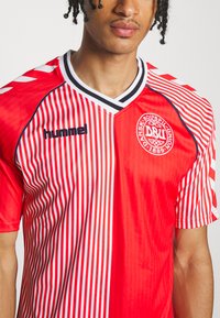 Nuori mies, joka pukeutunut puna-valkoiseen Tanskan jalkapallopaitaan, jossa on hummelin logo ja Tanskan jalkapalloliiton vaakuna.