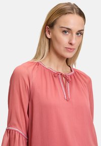 Blusa rosa con scollo rotondo, nodo frontale e delicate rifiniture. Maniche lunghe con polsini a campana e tessuto leggero e textured.