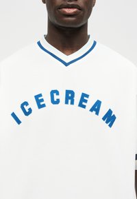 Μαύρο άτομο που φορά λευκό πουκάμισο με μπλε περιθώριο και τη λέξη «ICECREAM» κεντημένη με μπλε στο στήθος.
