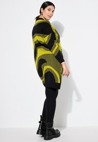 Maglione oversized in nero e verde lime con motivi ondulati, abbinato a leggings neri e stivali neri chunky. Tessuto testurizzato, vestibilità rilassata.