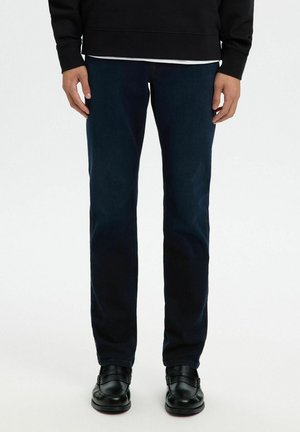 Mand iført mørkeblå jeans med lige ben, sorte læderloafer og sort langærmet top, står med afslappede hænder ned langs siden.