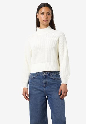 Kvinne iført en hvit ribbestrikket genser og høyreiste blå jeans, som ser litt til siden mot en ensfarget bakgrunn.