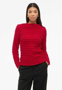 Rød tekstureret sweater med et gittermønster, rund halsudskæring og lange ærmer, parret med sorte bukser. Blødt materiale tilføjer dybde.