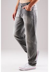 Graue Jeans aus Denim mit lockerer Passform, ausgestattet mit Fronttaschen, einem Reißverschluss und kupferfarbenen Akzenten. Kombiniert mit weißen Sneakern.