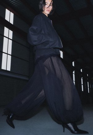 Nő beltéren sétál fekete bomber dzsekit, áttetsző bő szárú nadrágot és fekete, hegyes orrú, magas sarkú csizmát viselve gyenge fényben.