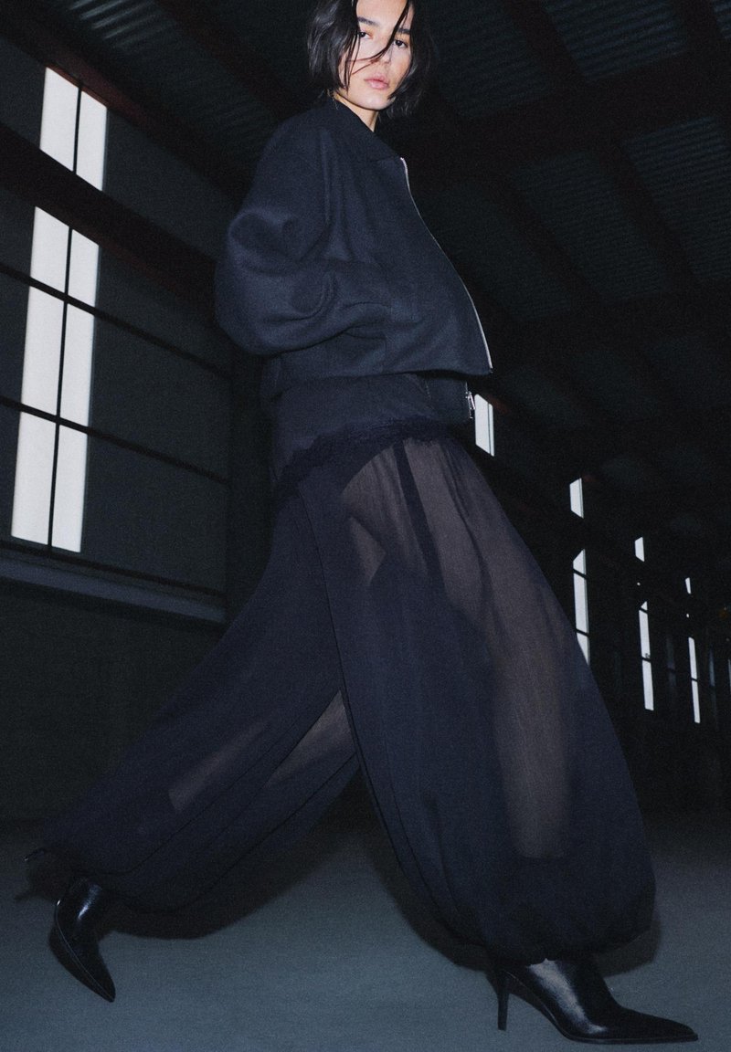 Frau läuft drinnen und trägt eine schwarze Bomberjacke, transparente Hosen mit weiter Beinweite und schwarze spitze Stiefel mit Absatz bei schwachem Licht.