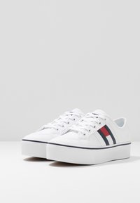 Sneaker bianchi in tela con punta rotonda, suola bianca spessa e dettagli in navy. Presentano strisce rosse e navy sui lati. Superficie texturizzata.