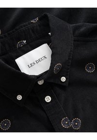 Camicia di velluto a coste nera con bottoni bianchi, motivi geometrici ricamati in blu e marrone, e un'etichetta del marchio visibile.