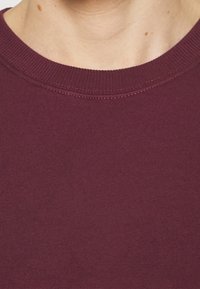 Sweter z dzianiny w kolorze bordowym, z prążkowanym, okrągłym dekoltem, o gładkiej fakturze i równych szwach. Brak widocznych wzorów lub metalowych elementów.