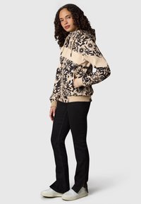 Femme portant un sweat à capuche zippé beige et noir à motif floral, pantalon noir avec fentes latérales et baskets blanches, debout devant un fond gris.