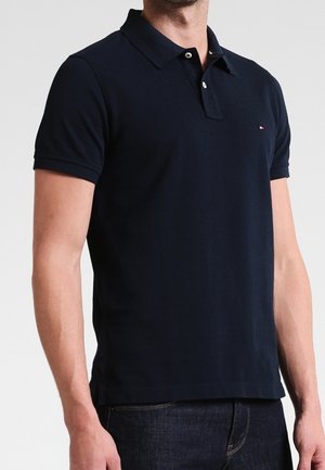 Mand iført en tætsiddende mørkeblå polo med korte ærmer, knappet krave og lille logo på venstre bryst, kombineret med mørke jeans.