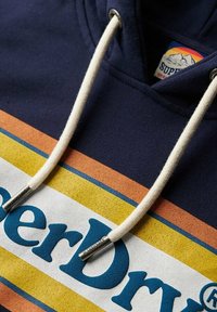 Granatowy hoodie z białą taśmą i niebieskim napisem "Superdry". Zawiera pasek w kolorowe paski w odcieniach pomarańczowym, żółtym i niebieskim. Wykonany z materiału bawełnianego.