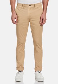 Beige katoenen chinos met een slanke pasvorm, voorzien van een knoopsluiting, zijzakken en opgerolde mouwen. Draagt met witte sneakers.