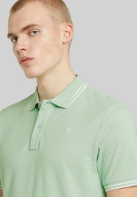 Grön polo t-shirt med texturerat tyg, tvåknappslå, och vita randiga krageaccent; har en liten logotyp på bröstet.