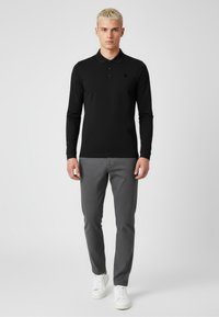 Schwarzes Langarm-Polohemd mit Kragen, ausgestattet mit drei Knöpfen, kombiniert mit grauen strukturierten Hosen und weißen Sneakers.
